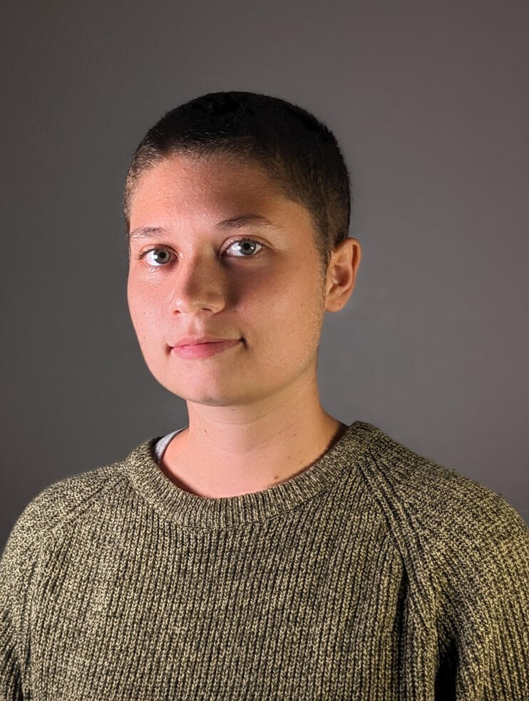 A portrait of Abi Walls. Headshot of a mixed race genderqueer person with short brown hair and green eyes against a grey background, wearing a green sweater.