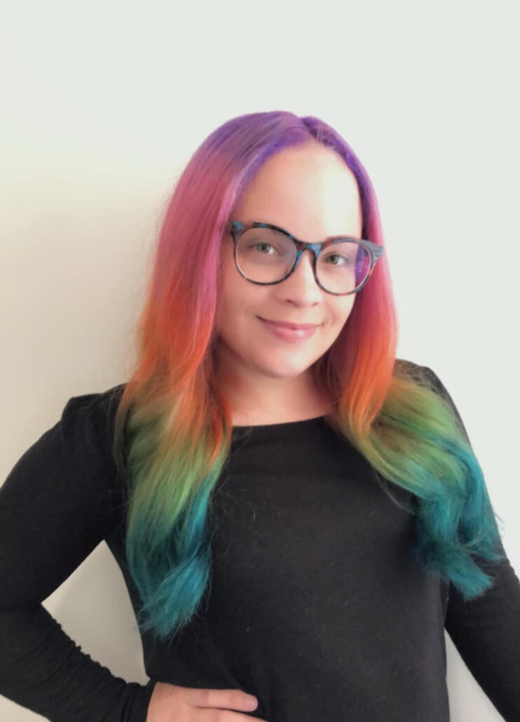 A smiling light-skin multiracial woman with long wavy rainbow hair and large round dark glasses in a black shirt standing in front of a white wall.